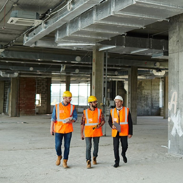 Group of three workmen wearing protective helmets and vests on construction site: walking among concrete walls at basement floor of unfinished building with foreman inspector, wide shot